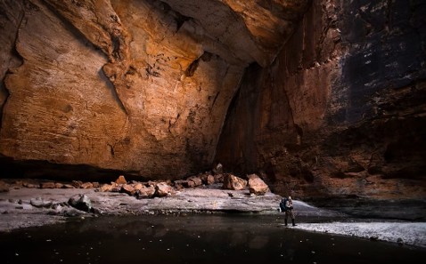 A lone figure stands in a vast ancient canyon, dwarfed by towering ochre walls